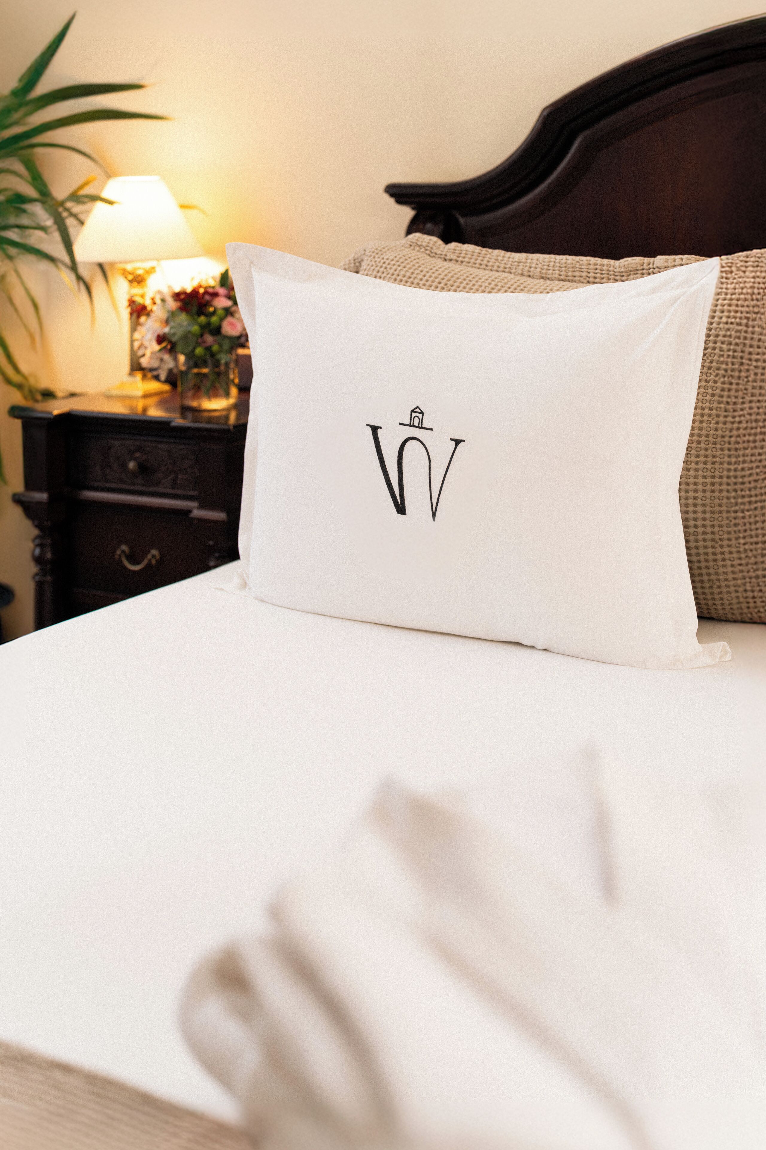 Cama bem arrumada com travesseiro branco com logotipo, cabeceira de madeira escura e mesa de cabeceira com abajur e flores.
