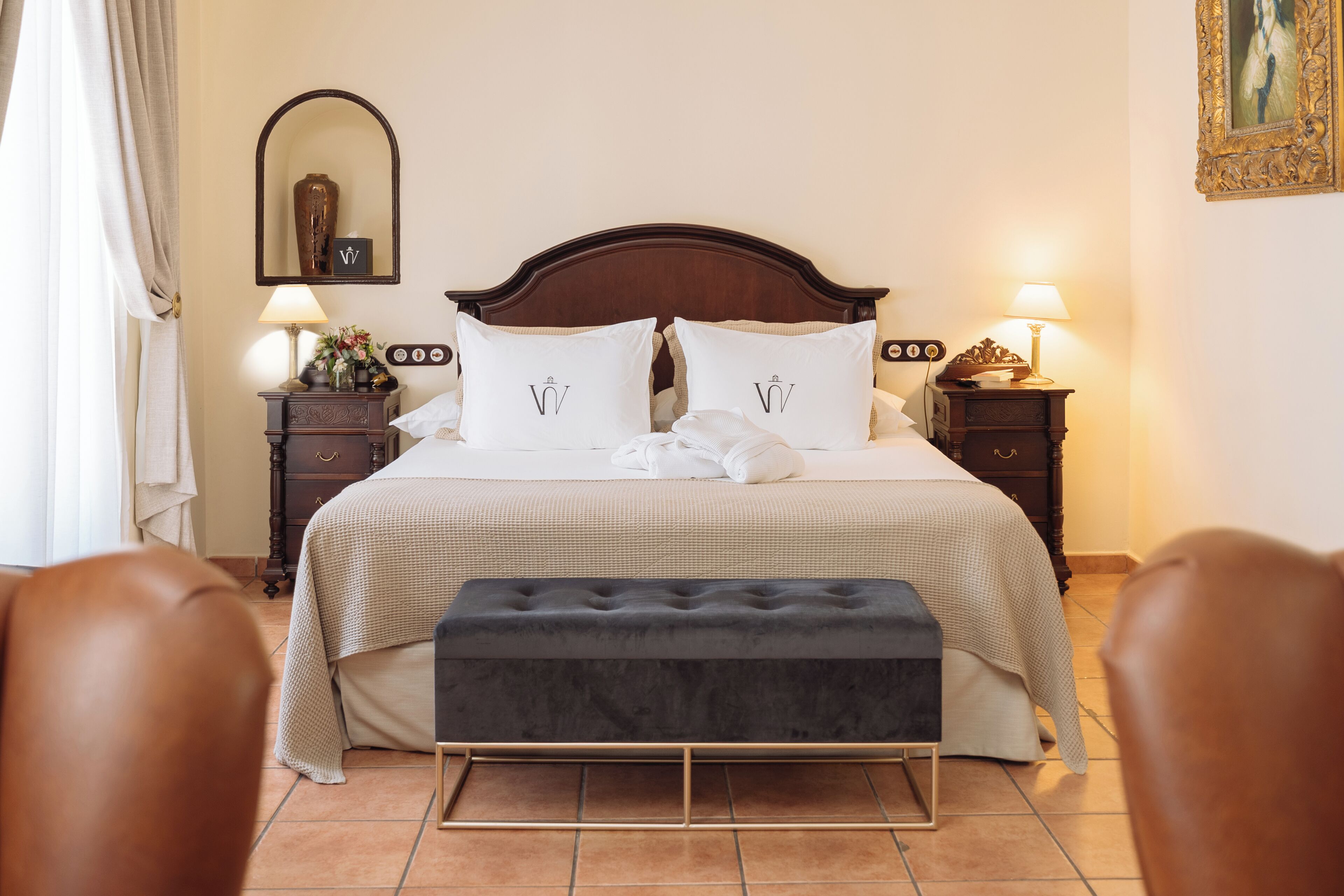Un lit bien fait, avec une tête de lit, deux tables de chevet avec des lampes, un banc et un miroir accroché au mur.