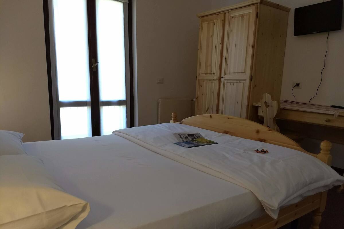 Un lit simple avec une tête de lit en bois, un matelas blanc et un magazine posé dessus. On aperçoit une armoire en bois à deux portes et un téléviseur fixé au mur, à droite.