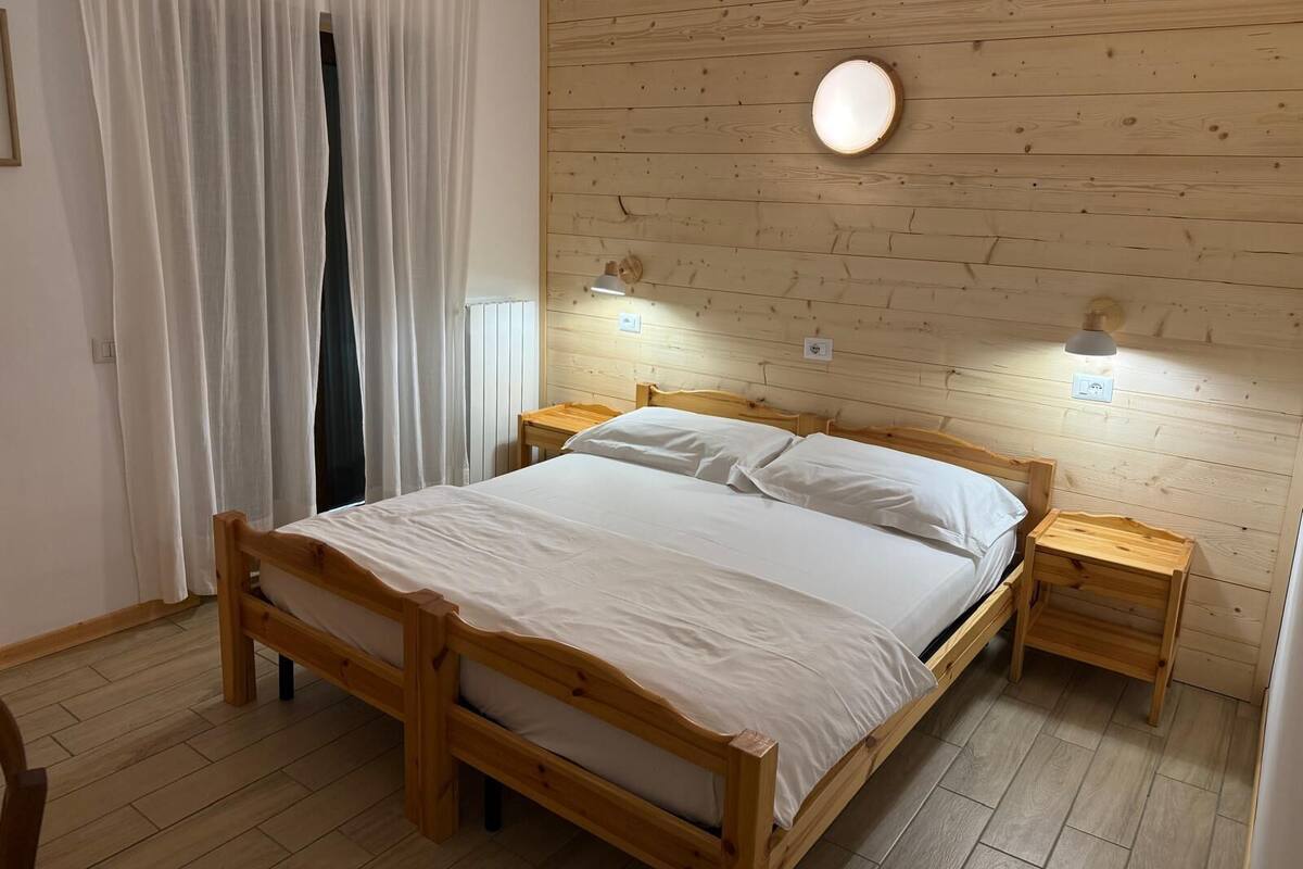 Un lit en bois avec une literie blanche, deux tables de chevet, une lampe fixée au mur et une fenêtre avec des rideaux.