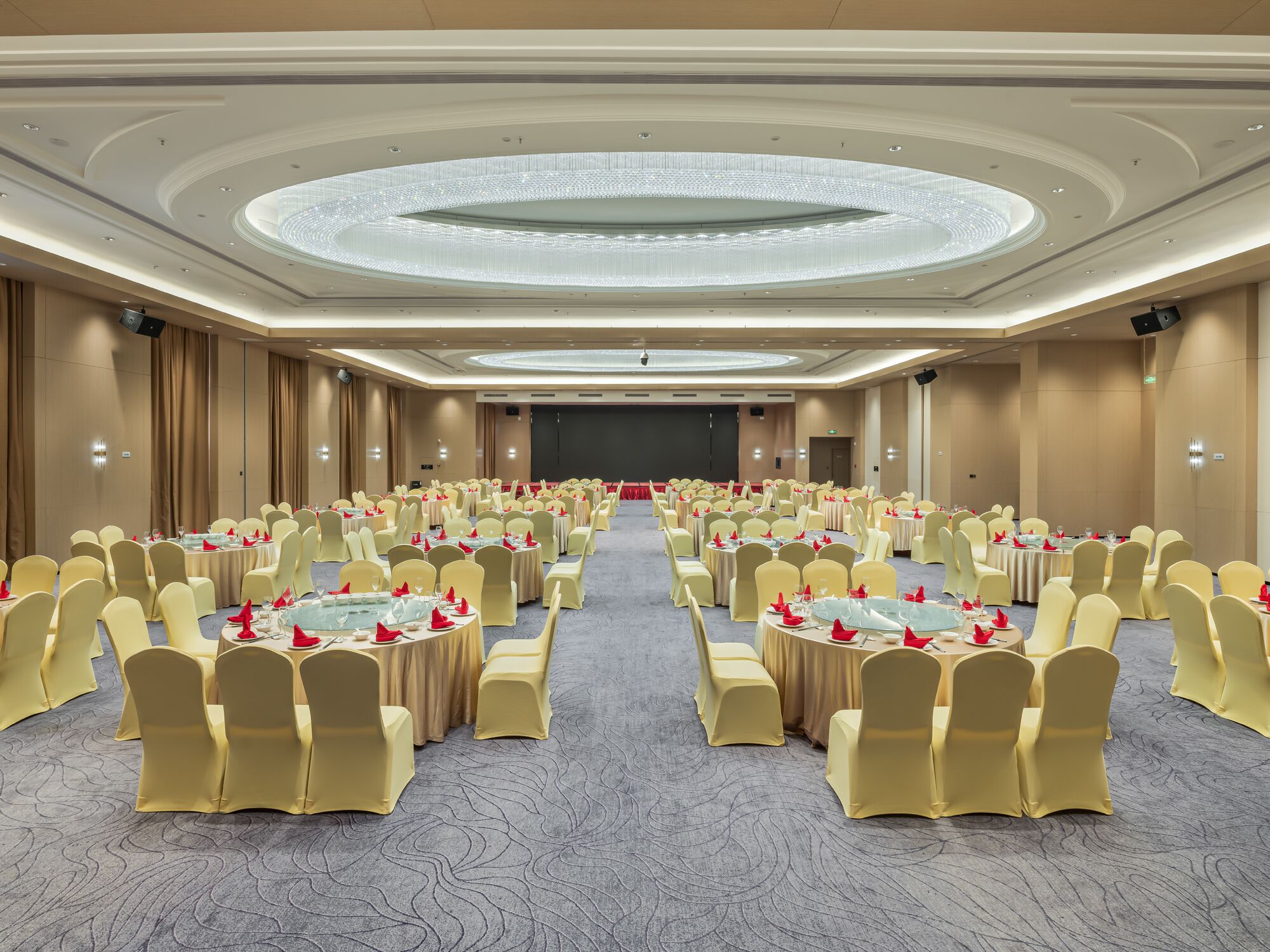 Une grande salle de conférence dotée de tables et de chaises rondes, d’une scène centrale et d’un plafond haut surmonté d’un luminaire circulaire.