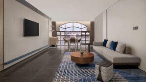 A modern hotel room with a large window, a flat-screen TV, a sofa, a coffee table, and a patterned rug.