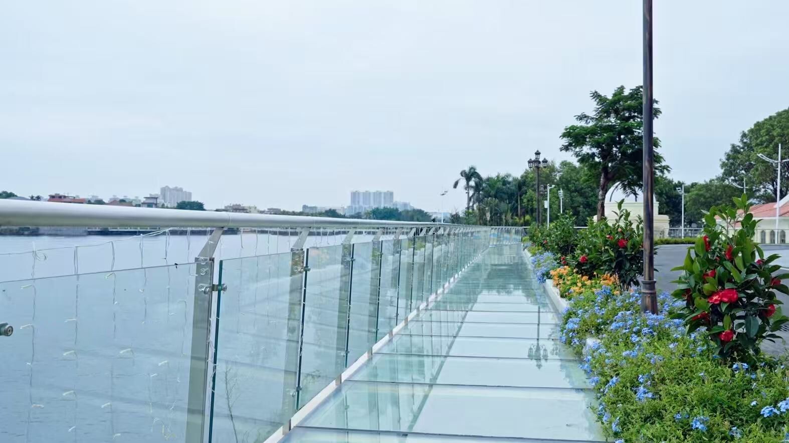 A glass bridge with a metal railing, a river, and a row of flowers and plants on the right.