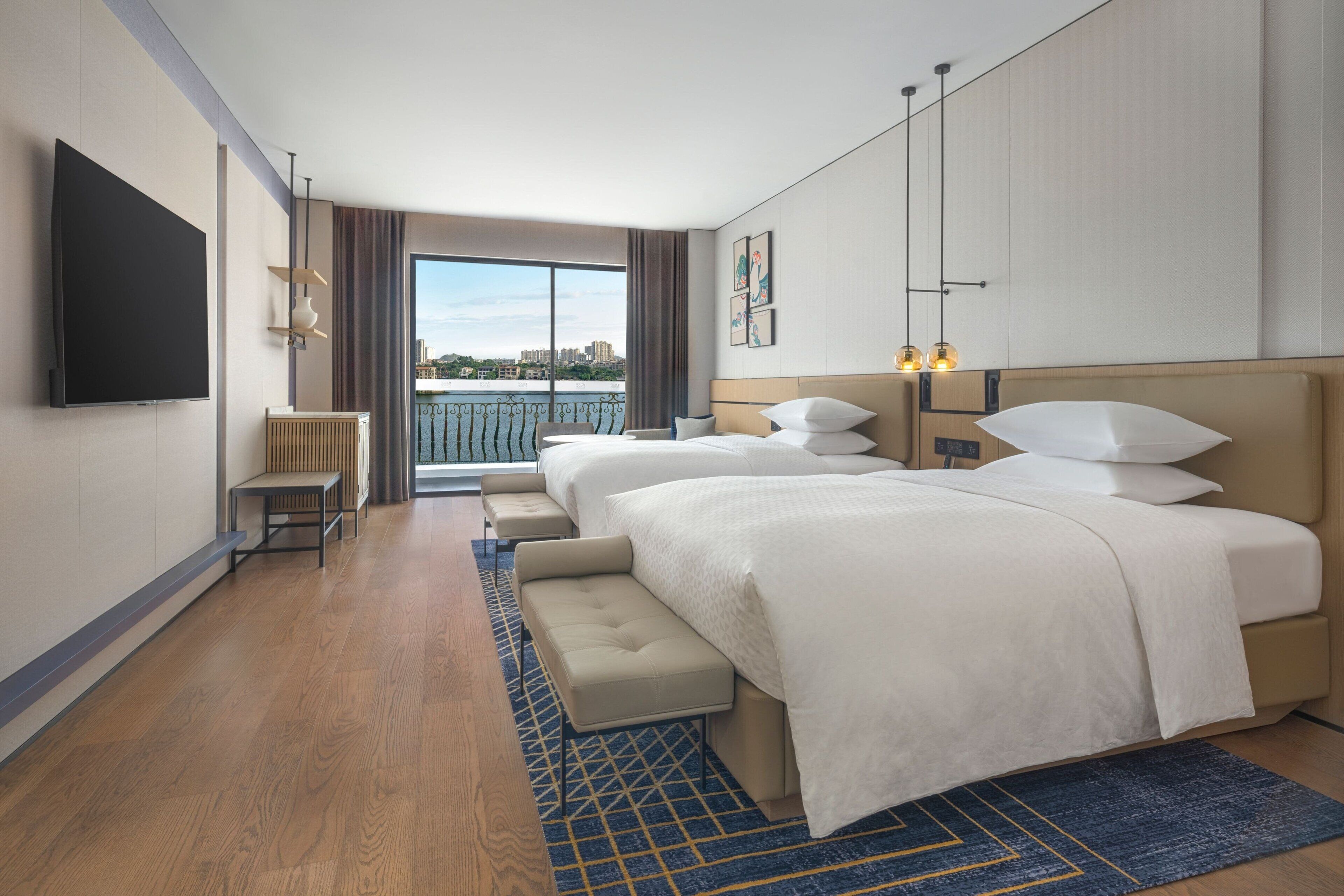 A hotel room with two beds, a TV, a bench, and a view of the city.