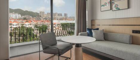 A modern hotel room with a large window offering a city view, a grey sofa, a round table, and a chair.