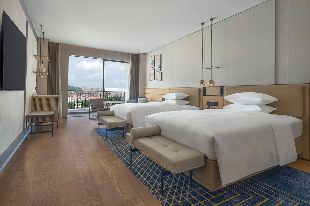 Une chambre d’hôtel avec deux lits, un coin salon, une grande fenêtre donnant sur la ville et un téléviseur à écran plat fixé au mur.