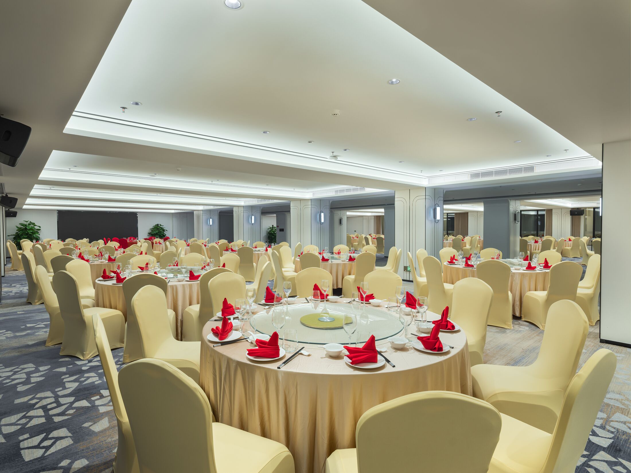 A large banquet hall with round tables, beige chairs, and red napkins.