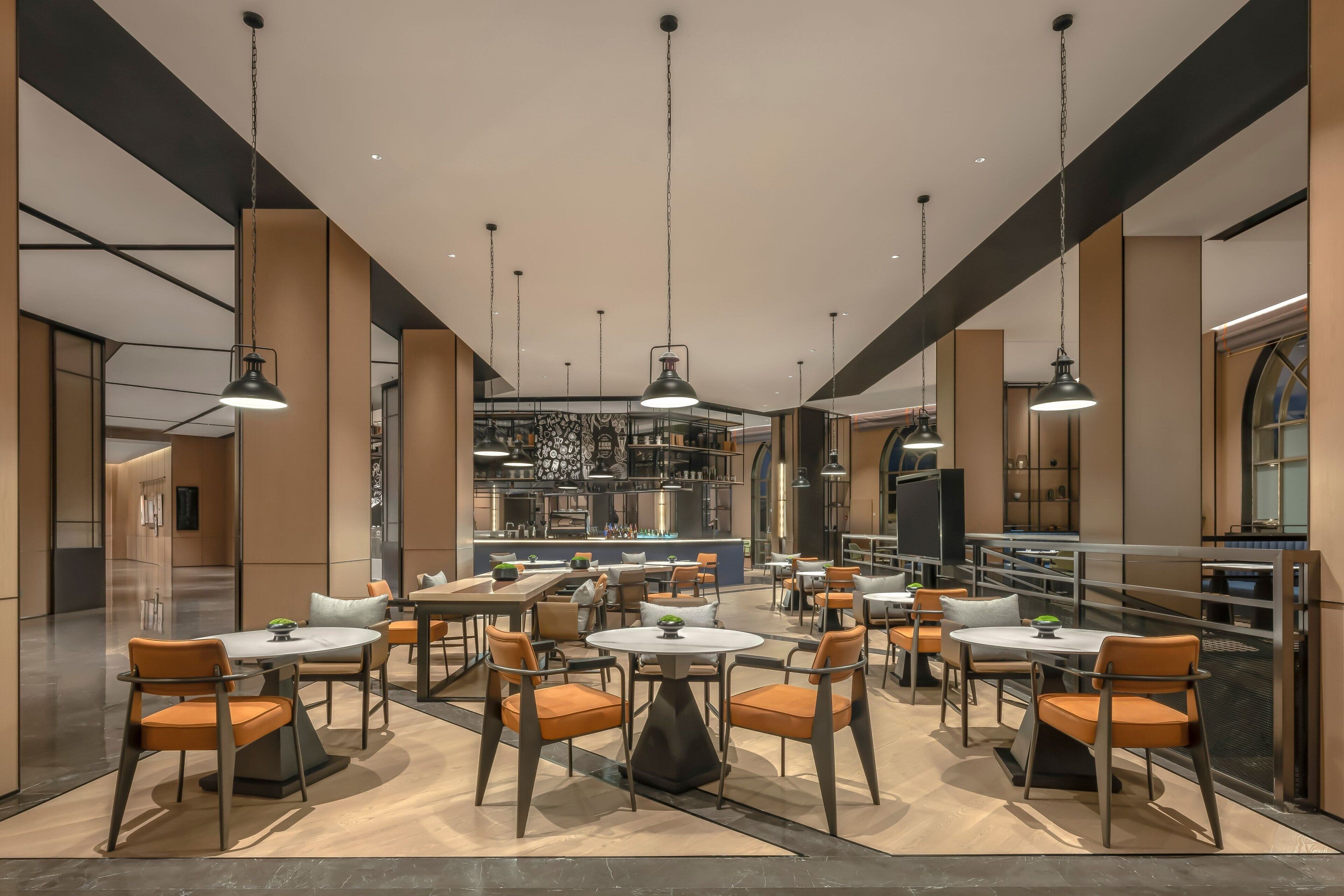 Un área de comedor moderna con mesas redondas y sillas naranjas, una cocina al fondo y vistas de la ciudad a través de grandes ventanales.