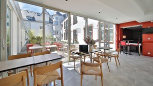 A spacious indoor area with large windows, a dining area with tables and chairs, and a kitchen area with red accents.