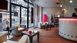 A modern hotel lobby with a reception desk, seating area, and large windows offering a view of the street.