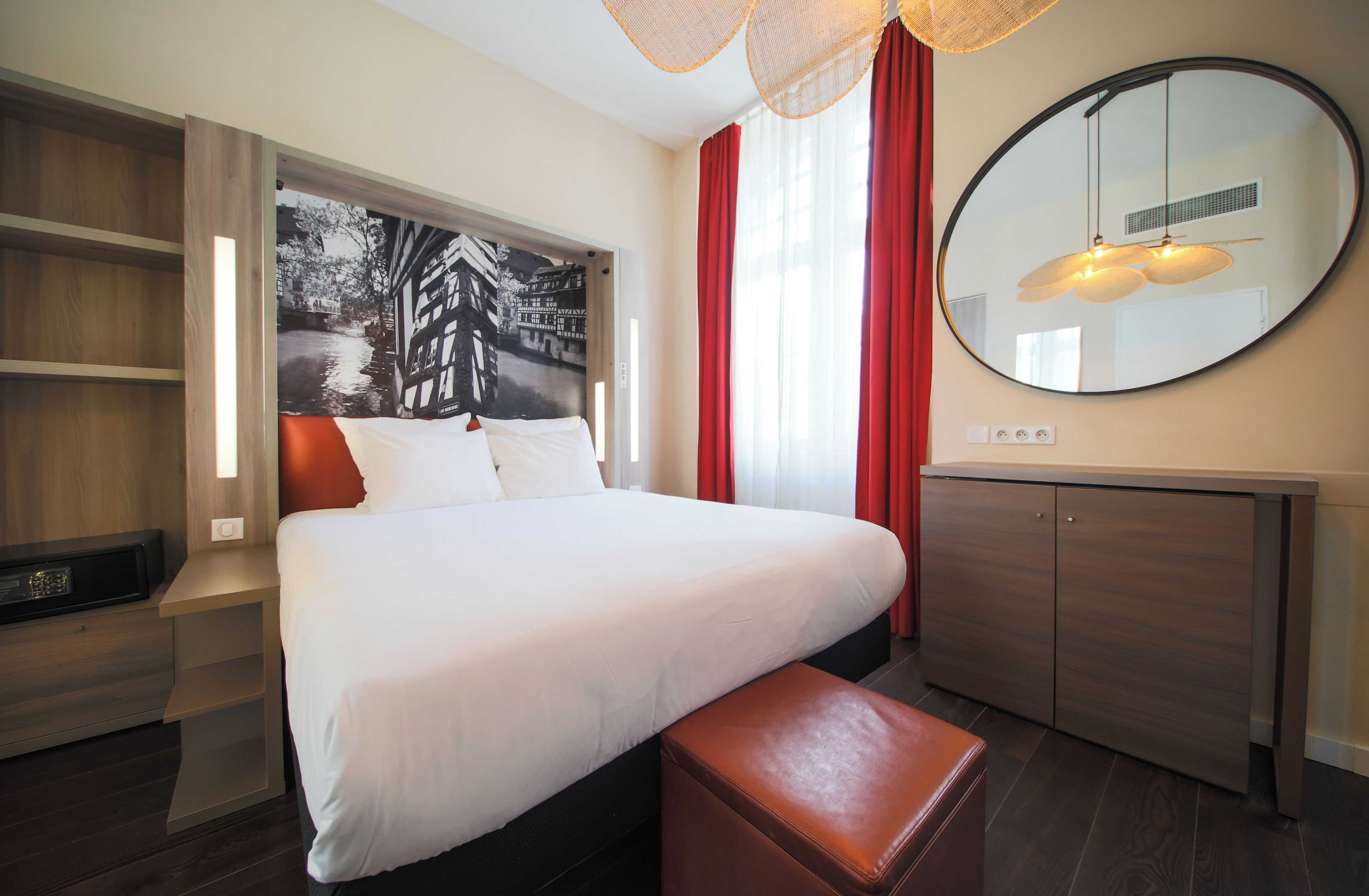 A modern hotel room with a large bed, a wooden nightstand, a bench, a wardrobe, a round mirror, and a wall-mounted light fixture.