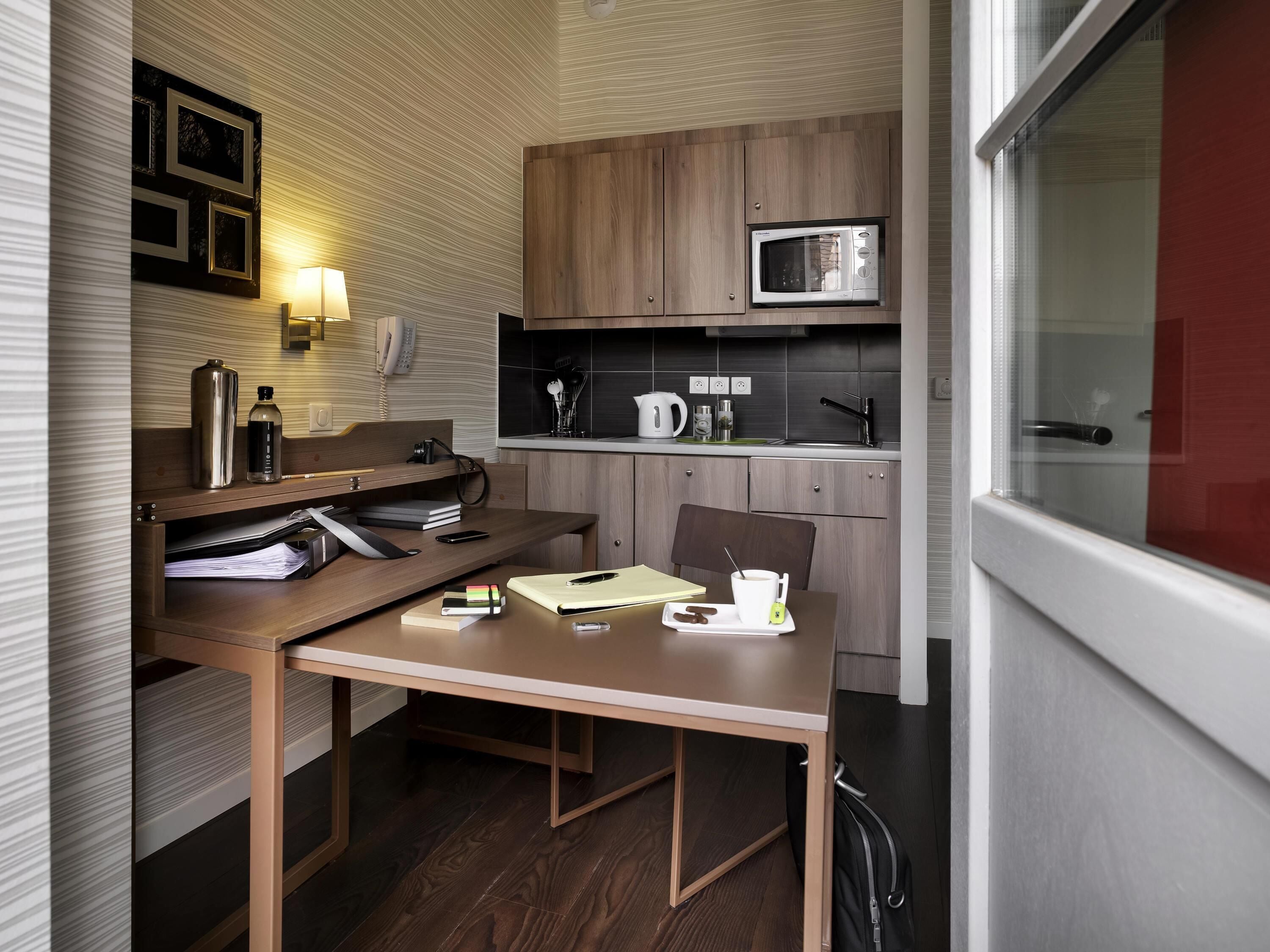 A compact kitchen with a microwave, kettle, and coffee maker. There is a wooden table with a chair, a small shelf with kitchen items, and a window with a view.