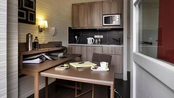 A compact kitchen with a microwave, kettle, and coffee maker. There is a wooden table with a chair, a small shelf with kitchen items, and a window with a view.