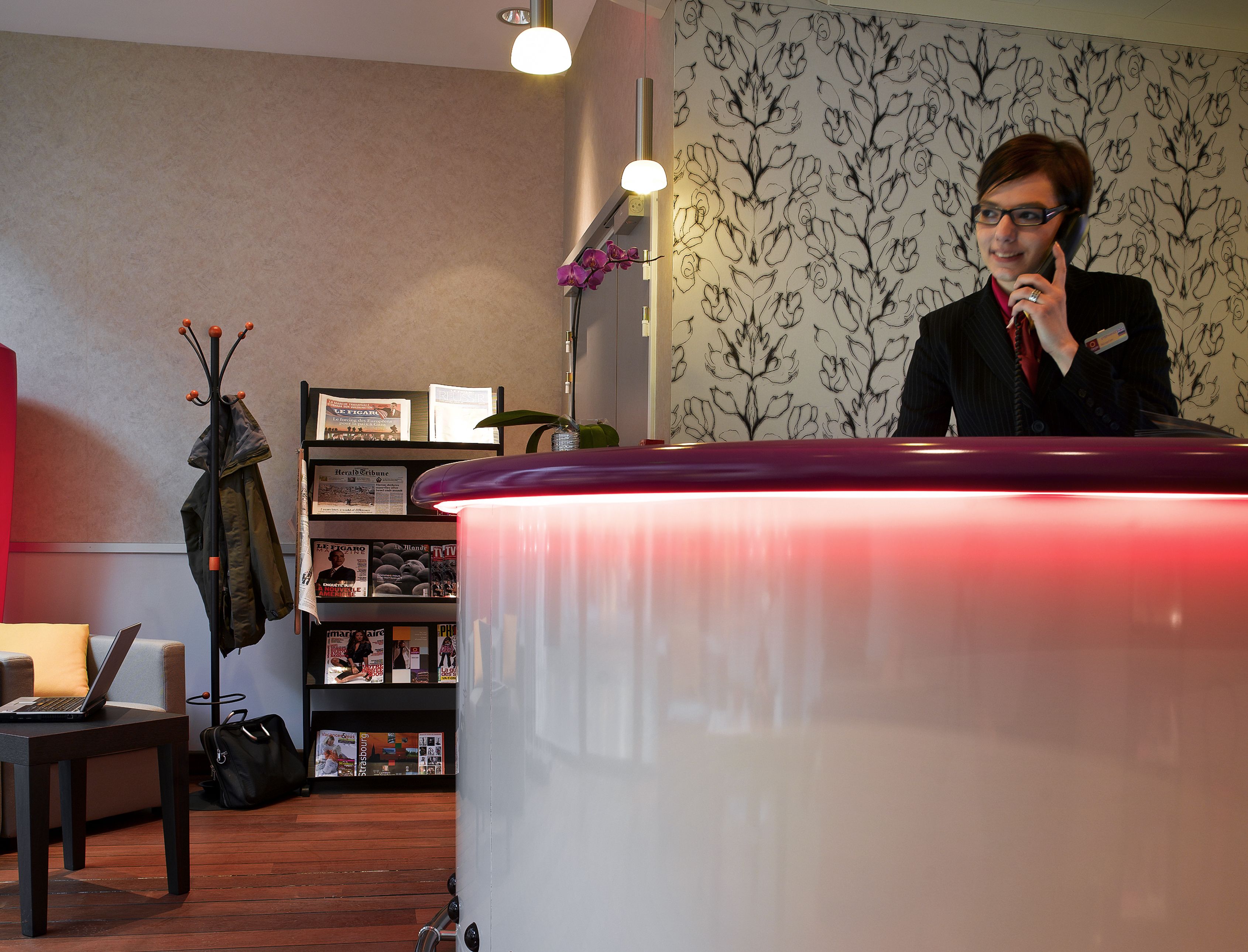 A reception desk with a red top, a laptop on a table, and a coat rack with jackets.