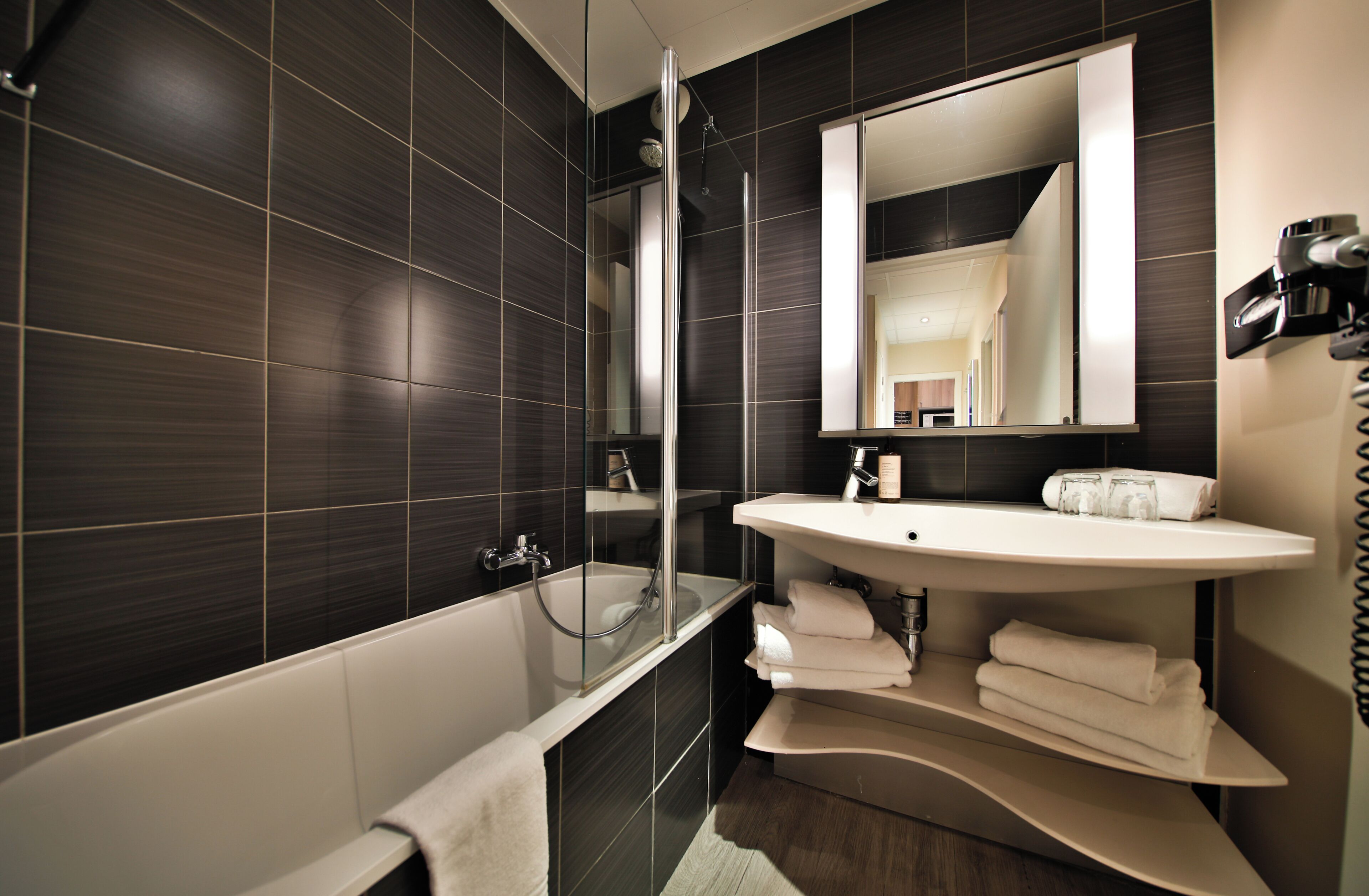 A modern bathroom with dark tiled walls, a glass-enclosed shower, a white sink, and a large mirror.