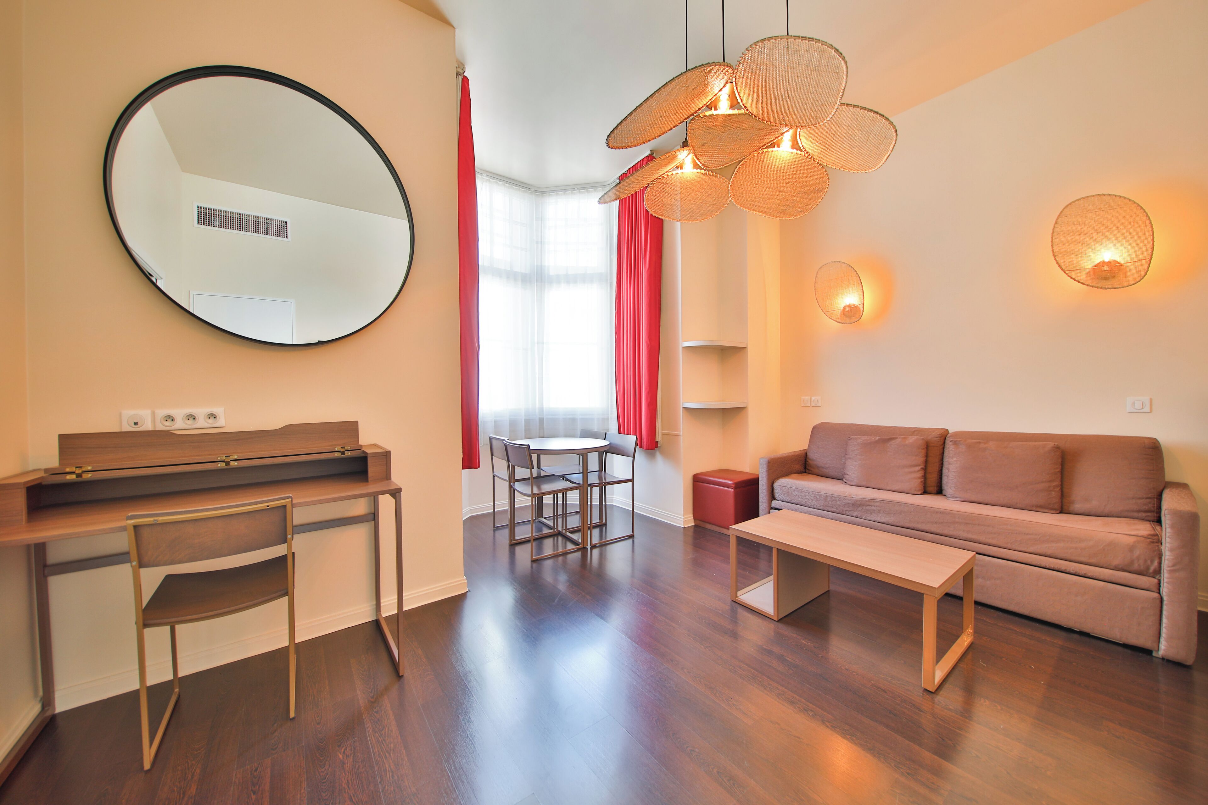 A modern living room with a sofa, a dining table, and chairs. There is a large mirror on the wall and a hanging light fixture.