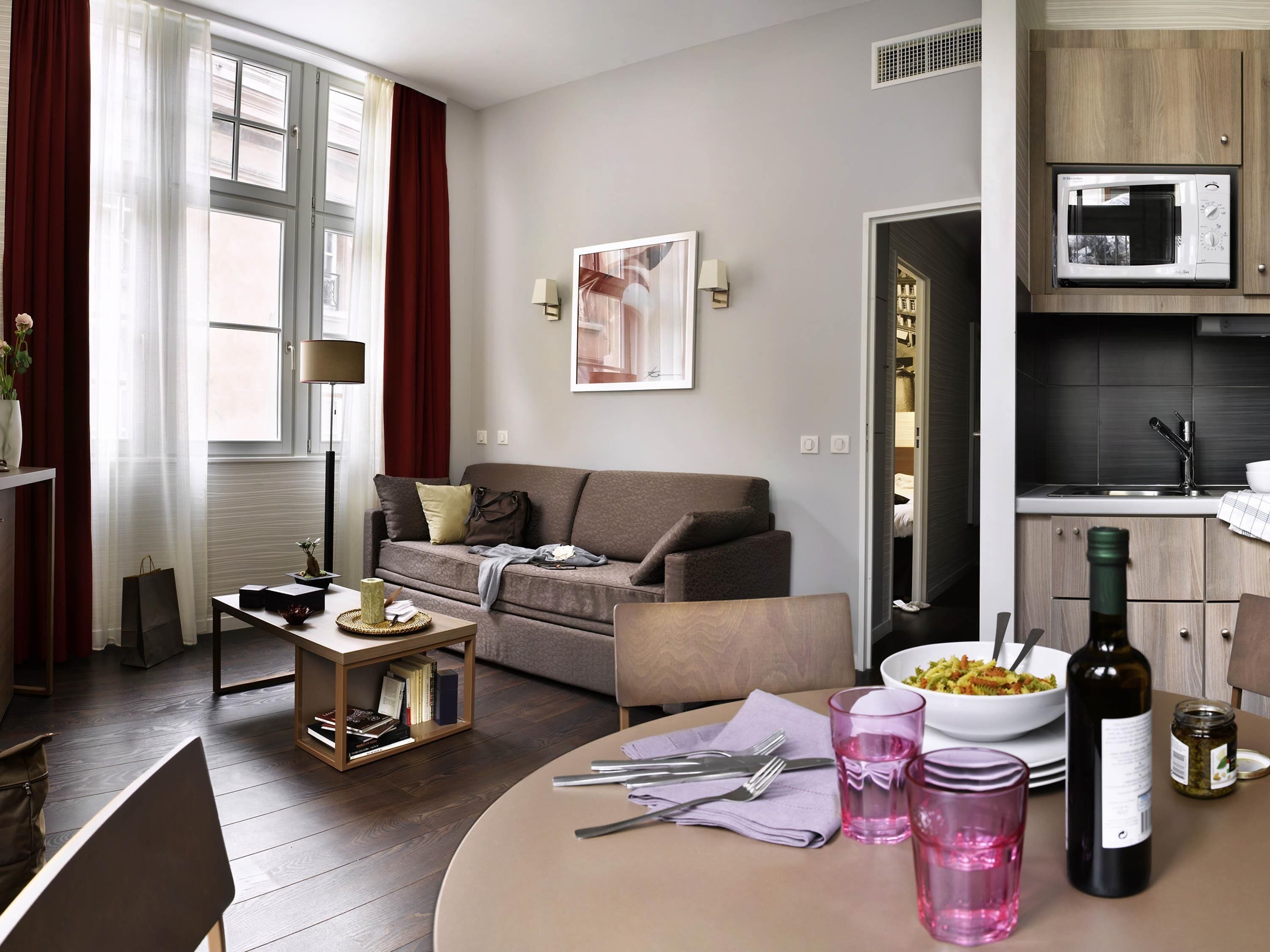 A modern living room with a sofa, a dining table with cutlery, a kitchen area with a microwave, and a bottle of wine.