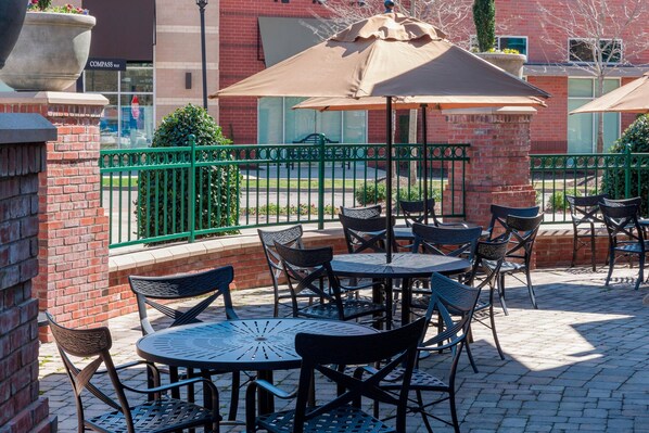 An outdoor patio with round tables and chairs, a green fence, and a brick building in the background.