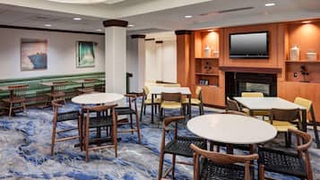 A hotel lobby with green cushioned seating, round tables, and chairs, a flat-screen TV mounted above a fireplace, and artwork on the walls.