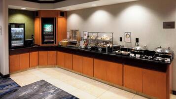 A hotel breakfast area with a buffet counter, refrigerated drinks, and various food stations.