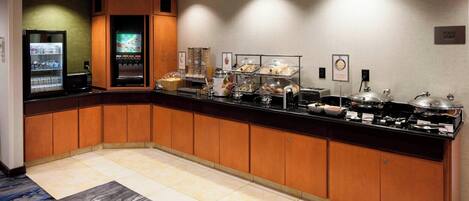 A hotel breakfast area with a buffet counter, refrigerated drinks, and various food stations.