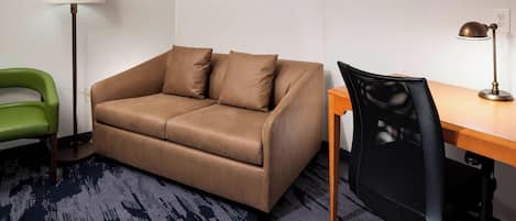 Una habitación con un sofá de cuero marrón, una silla verde, un escritorio de madera, una silla de oficina negra, una lámpara de pie y un cuadro enmarcado en la pared.