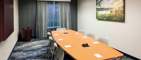 A conference room with a long table, chairs, a trash can, and a window with curtains.