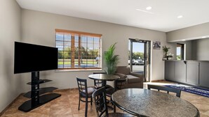 A modern hotel room with a flat-screen TV, a dining area with a round table and chairs, a potted plant, and a view of the parking lot through the window. - Americas Best Value Inn Byhalia (Byhalia)