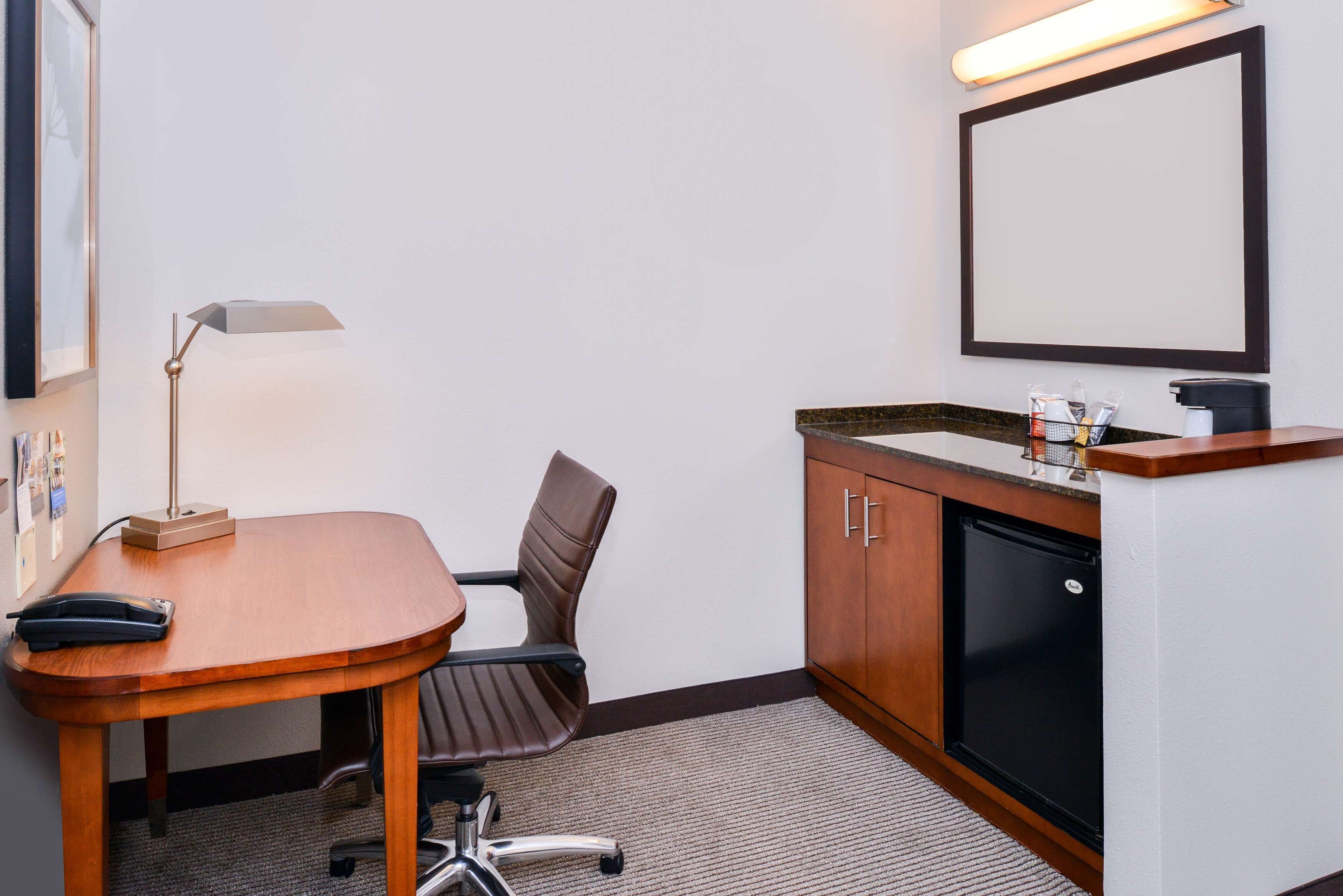 Un petit bureau avec un bureau en bois, une seule chaise de bureau, une lampe de bureau, un réfrigérateur et un miroir accroché au mur.