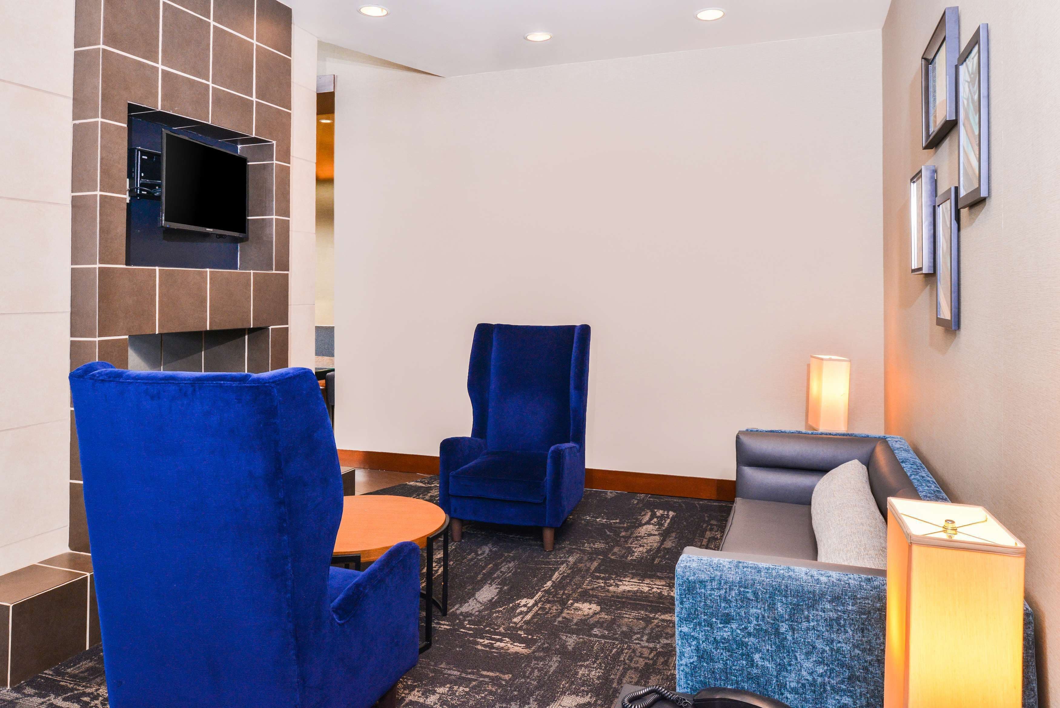 Eine Hotellobby mit einem an einer gefliesten Wand befestigten Flachbildfernseher, einem blauen Sessel, einem grauen Sofa und einem kleinen Tisch.