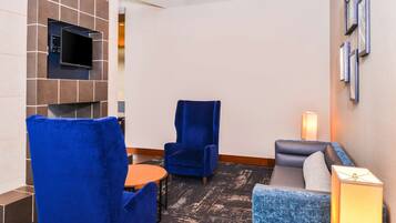 Eine Hotellobby mit einem an einer gefliesten Wand befestigten Flachbildfernseher, einem blauen Sessel, einem grauen Sofa und einem kleinen Tisch.