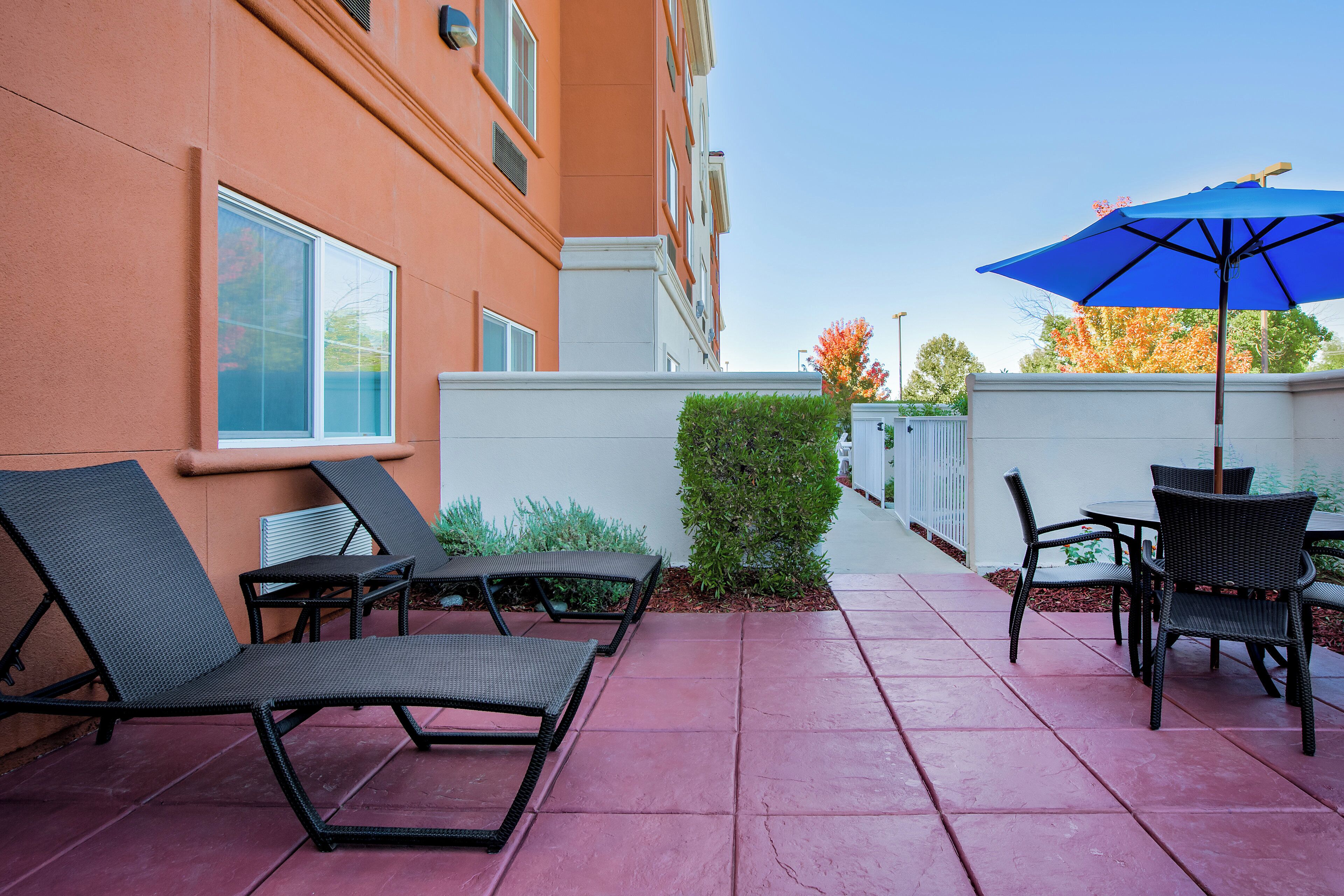 Una terrazza esterna con poltrone da lounge e un tavolo, un ombrellone blu, e un edificio dalle pareti arancioni e finestre.