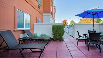 Una terrazza esterna con poltrone da lounge e un tavolo, un ombrellone blu, e un edificio dalle pareti arancioni e finestre.
