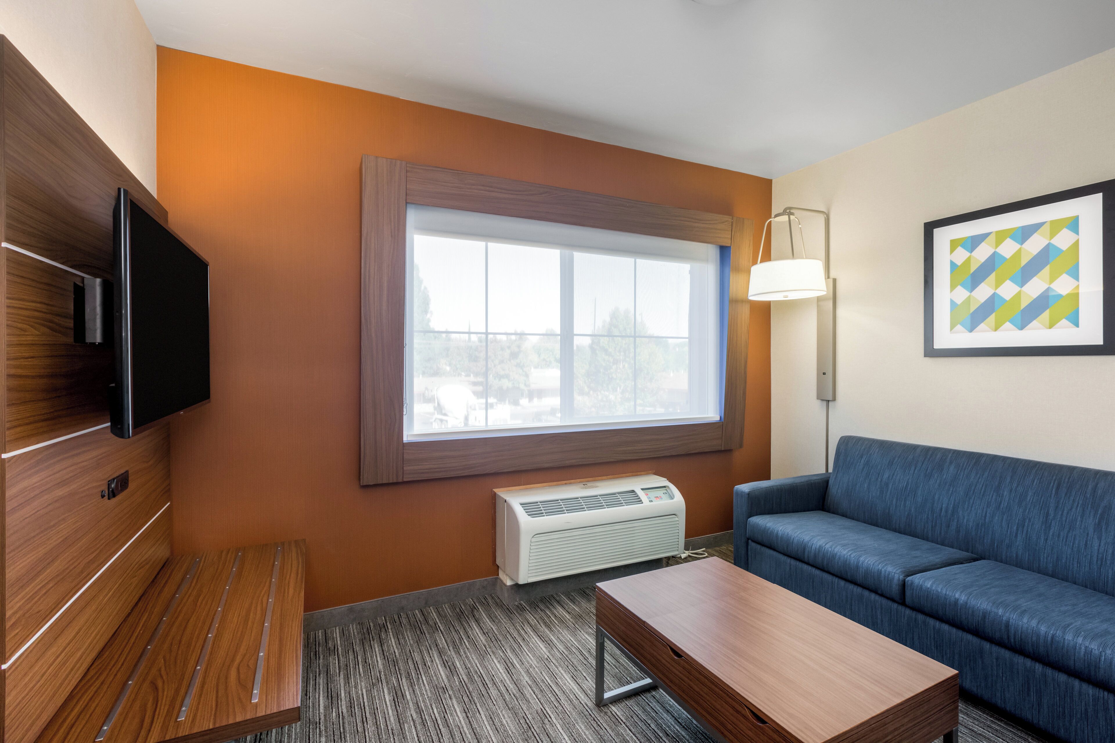 Een hotelkamer met een houten kast, een flatscreen-televisie, een raam met zonwering, een elektrische kachel, een blauwe bank en een ingelijst kunstwerk aan de muur.