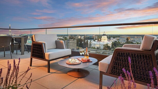 A rooftop terrace with a city view, furnished with a sofa, chairs, and a round table with a bottle and glasses.