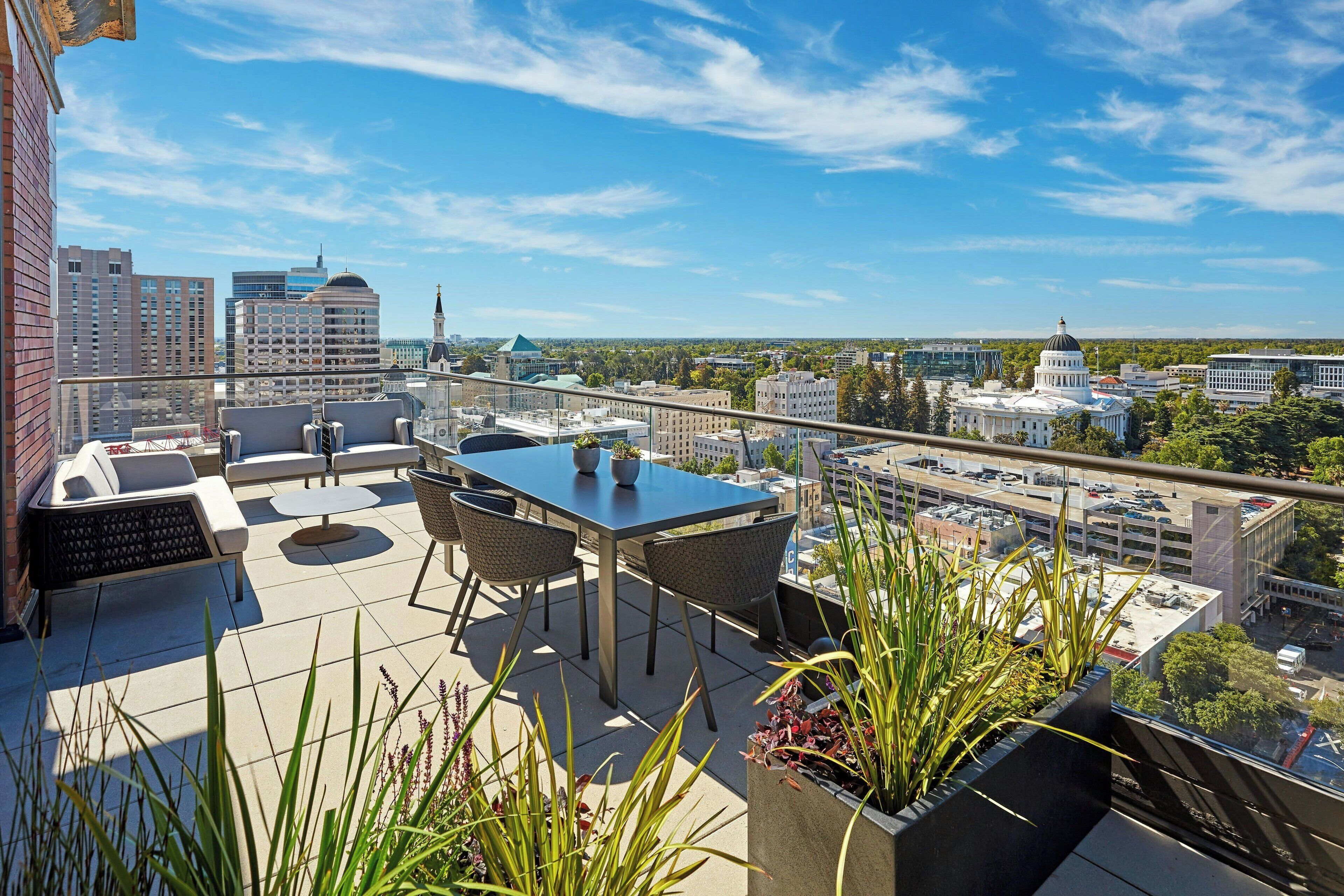 Una terraza en la azotea con mesa y sillas, rodeada de plantas en macetas, con vistas a un paisaje urbano lleno de edificios.
