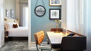 A hotel room with a bed, a desk with a chair, a lamp, and framed pictures on the wall.