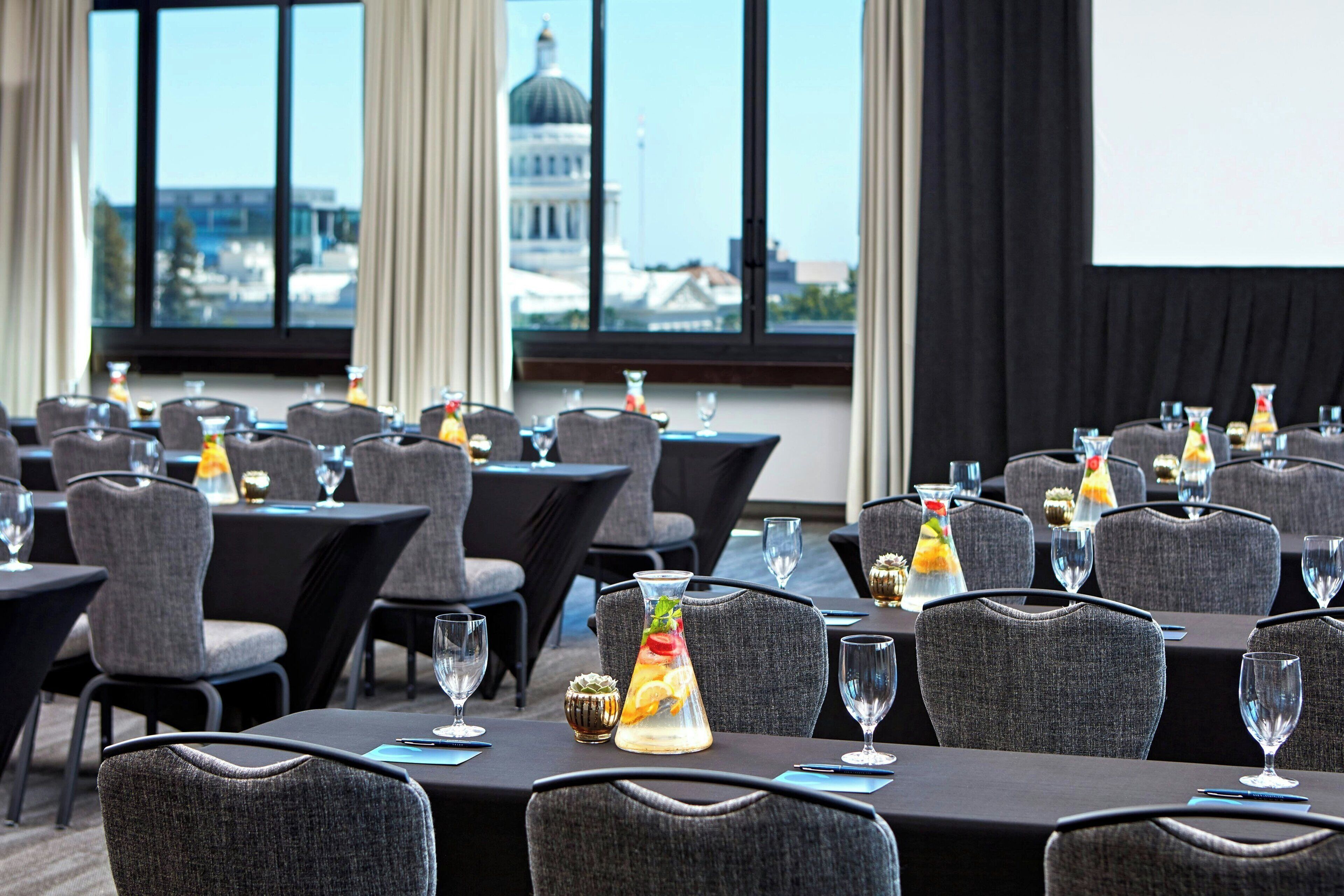 A conference room with tables set for a meeting, a projector screen, and a view of a building with a dome through large windows.