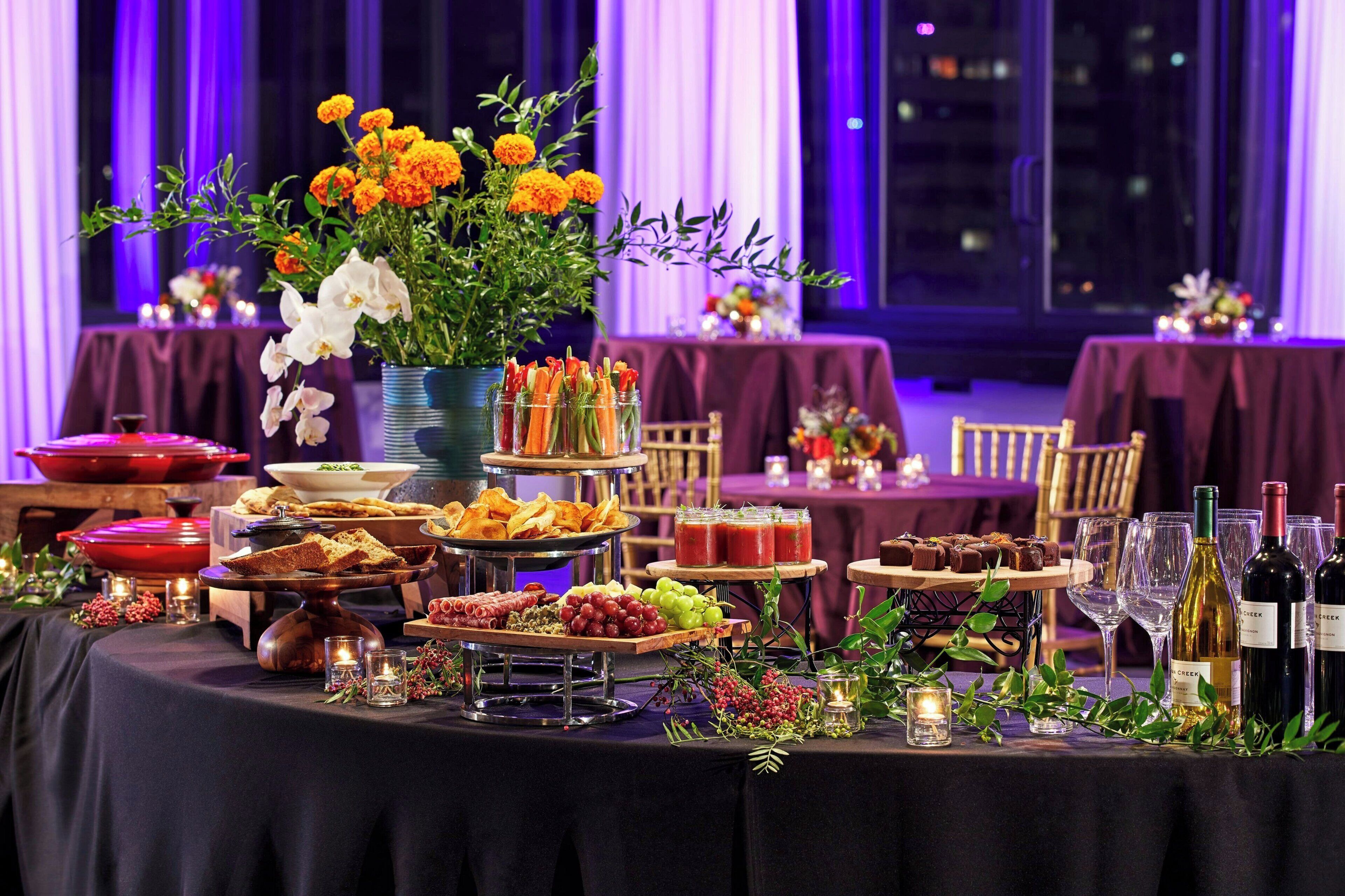 Una mesa de buffet con diversas opciones de comida y una selección de vinos, sobre un fondo de cortinas moradas y luces de la ciudad.