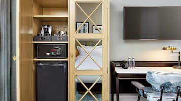 Una habitación de hotel compacta con minibar, cama con cabecera, televisor, escritorio con silla y una pequeña mesa con lámpara.
