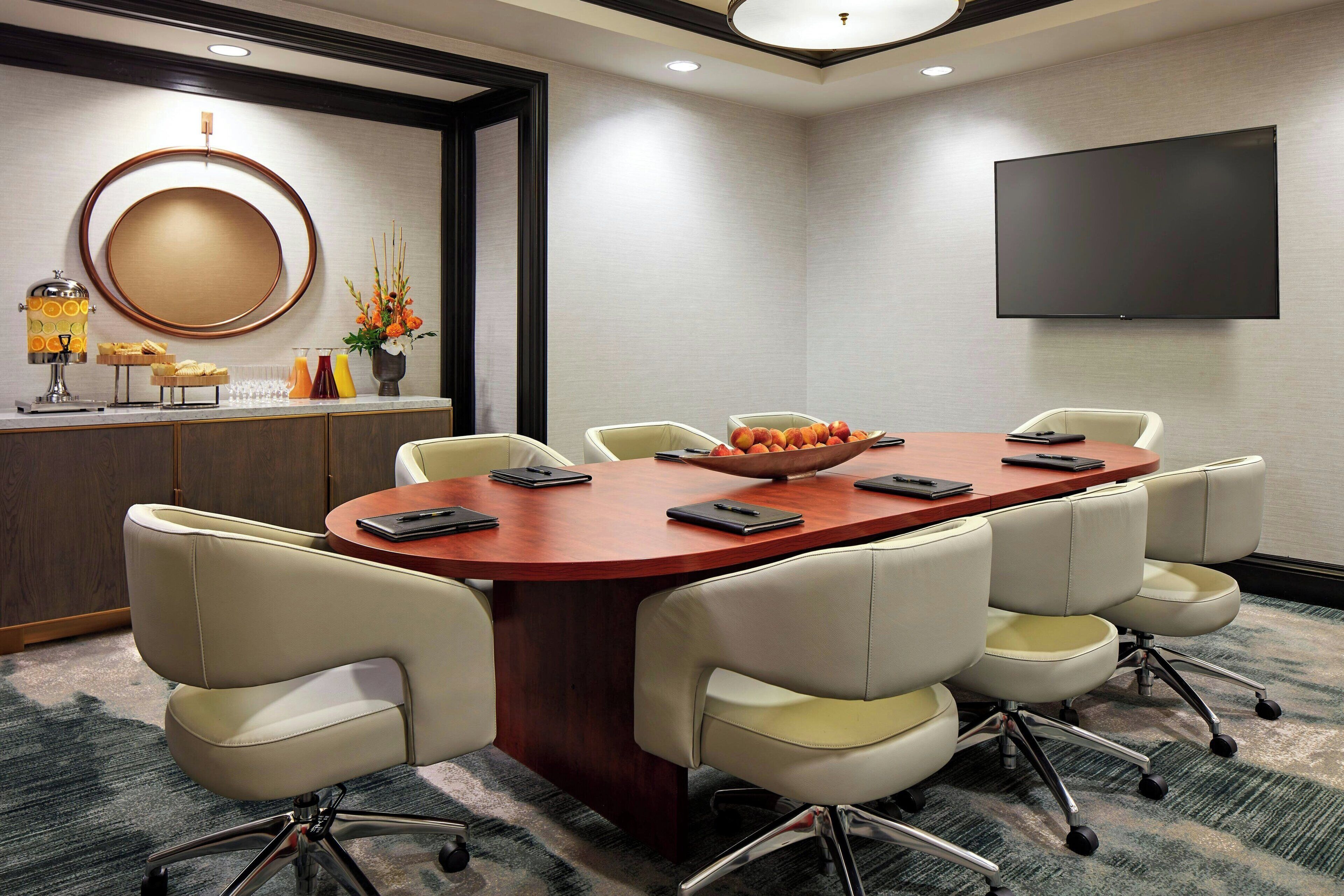 A conference room with a large oval table, six chairs, a TV, and a buffet area with fruit and drinks.