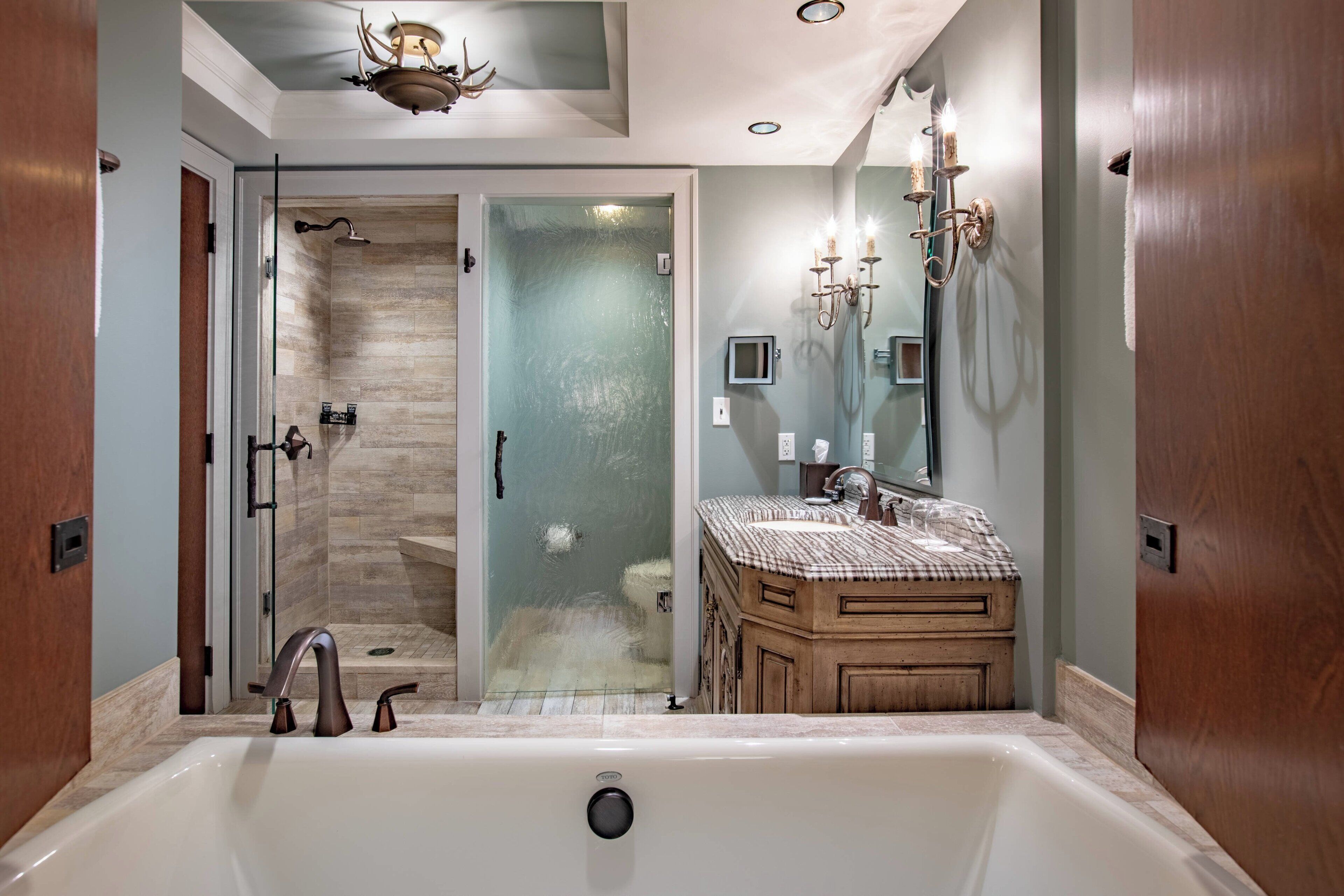 Une salle de bain moderne avec une grande baignoire, une douche à l’italienne avec des parois en verre, un meuble-lavabo en bois et un lustre.