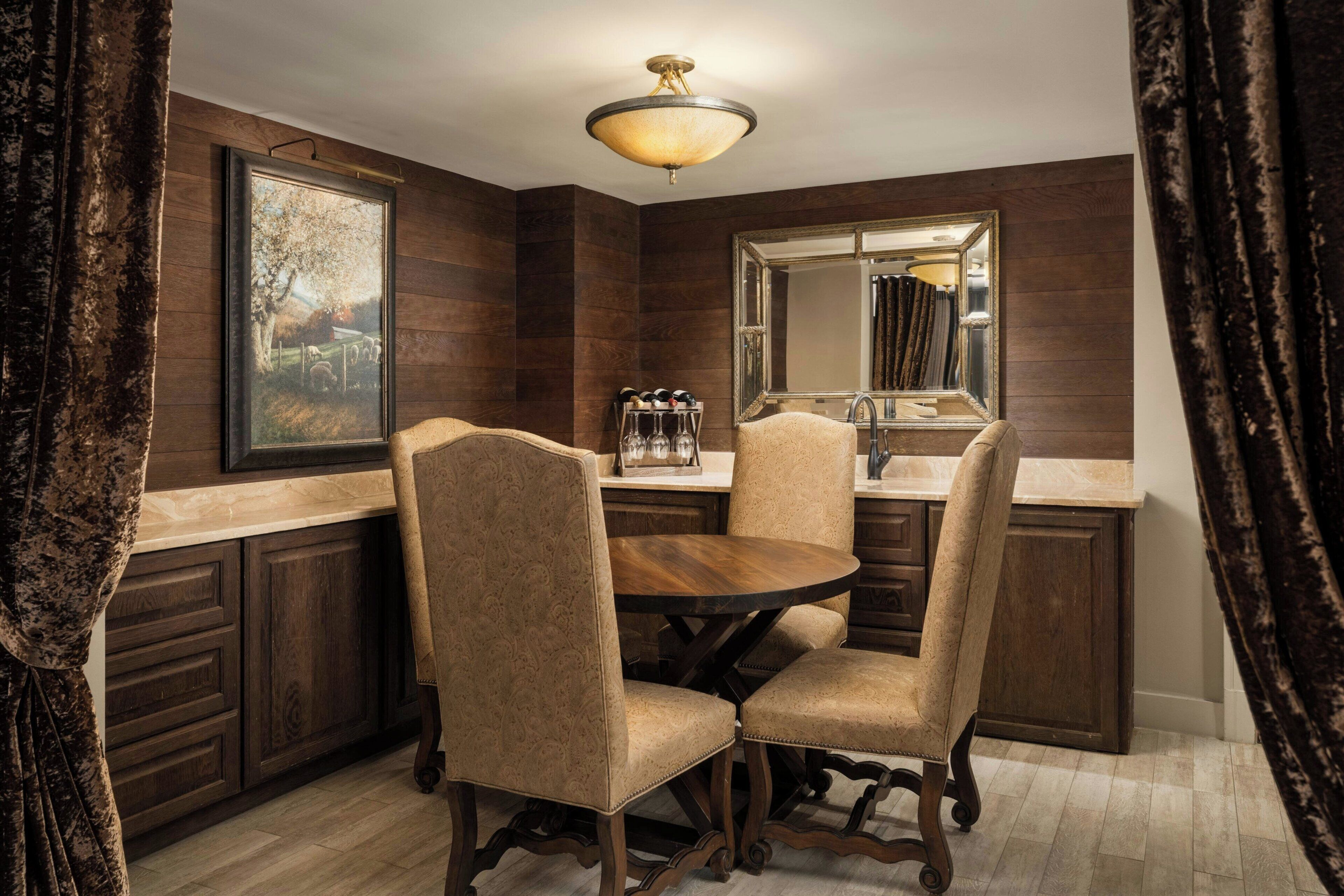 A dining area with a round wooden table, four upholstered chairs, a chandelier, and a large mirror above the sink.