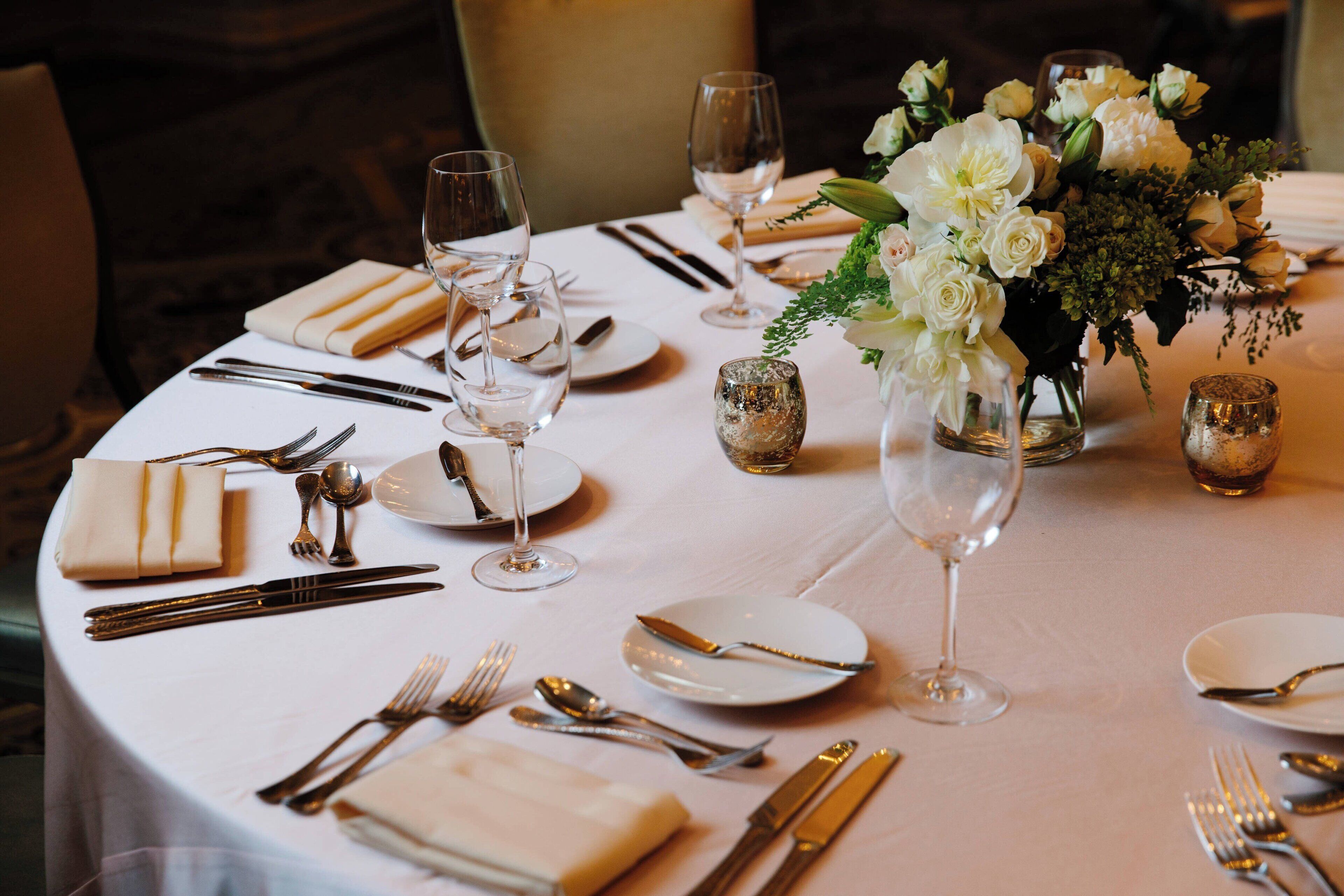Mesa redonda de comedor con mantel blanco, cubiertos, platos y copas de vino. Hay un florero con flores blancas y un pequeño adorno en la mesa.