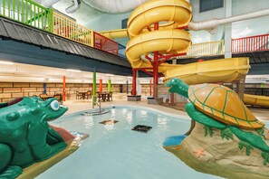 Parque acuático interior con tobogán, piscina con estructuras de juego de ranas y tortugas, y zona de descanso.