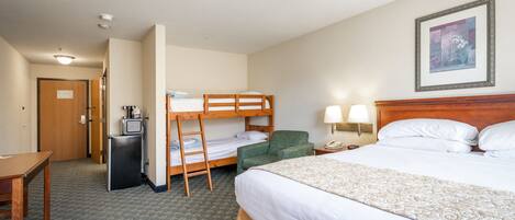A hotel room with a bunk bed, a desk, a small refrigerator, a bedside lamp, and a framed picture on the wall.