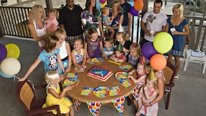 Un grupo de personas reunidas alrededor de una mesa con pastel de cumpleaños, globos y gorros de fiesta.