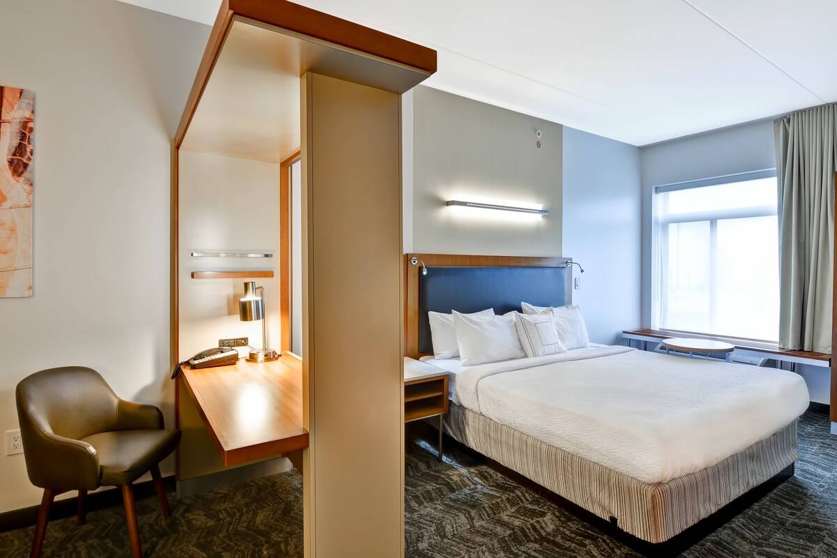 A hotel room with a large bed, a desk with a lamp, a chair, and a window with curtains.