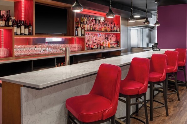 Een bar met een marmeren aanrecht, rode barstoelen en planken vol met verschillende flessen en glazen.