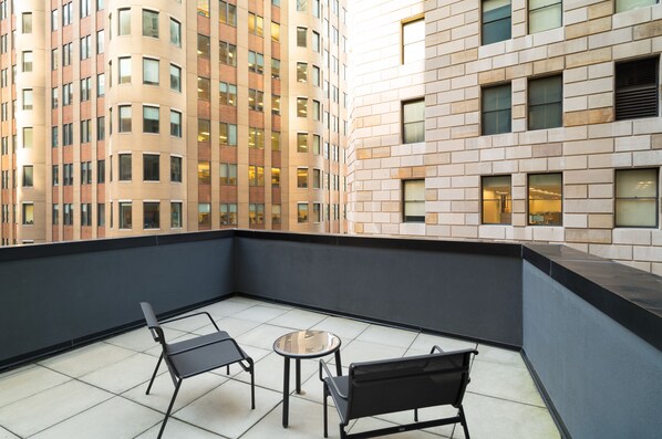 Un balcon avec deux chaises noires et une petite table, donnant sur un immeuble de grande hauteur à la façade en briques et aux nombreuses fenêtres.